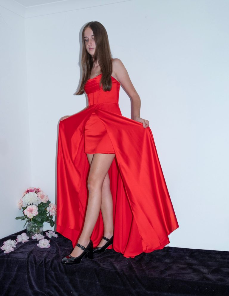 A woman in a long red dress with a high slit, standing on a dark surface with flowers nearby.
