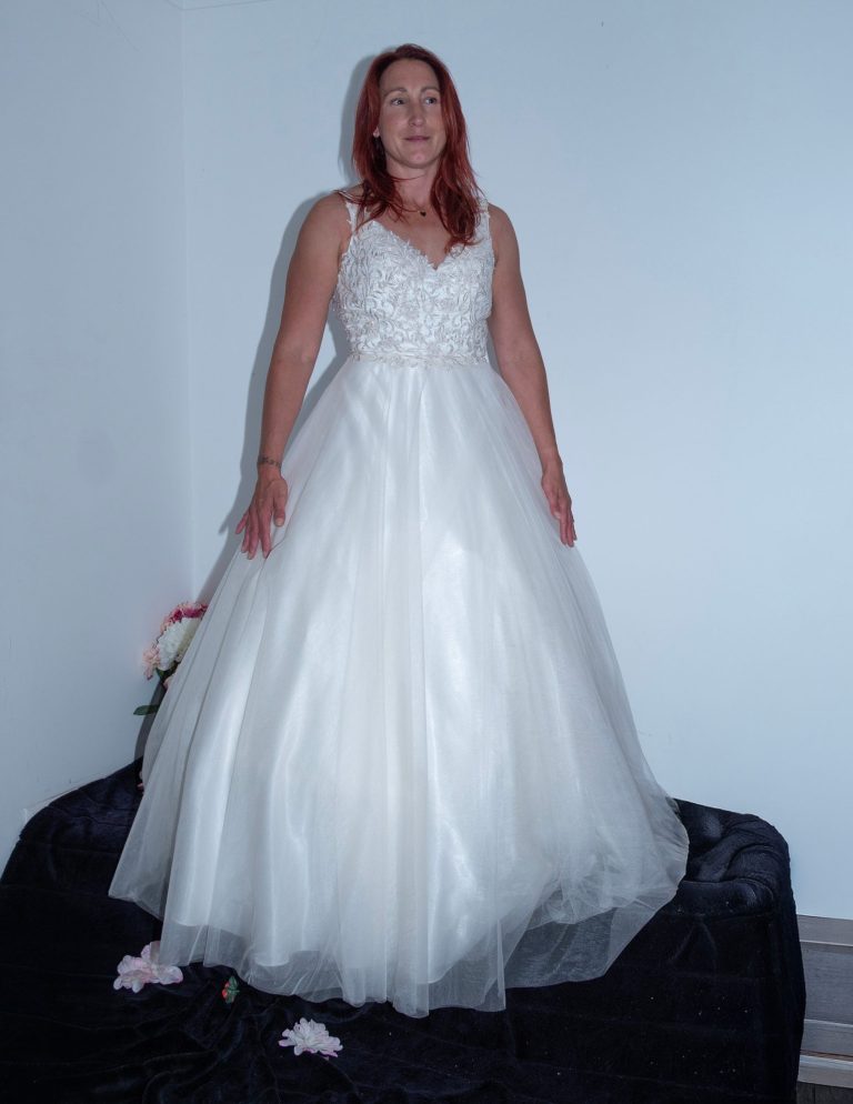 Woman in a white wedding dress, standing on a black surface with pink flowers nearby.