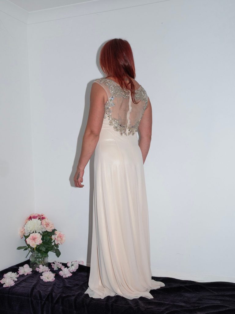 Woman in a long cream dress with detailed back, standing against a plain white wall.
