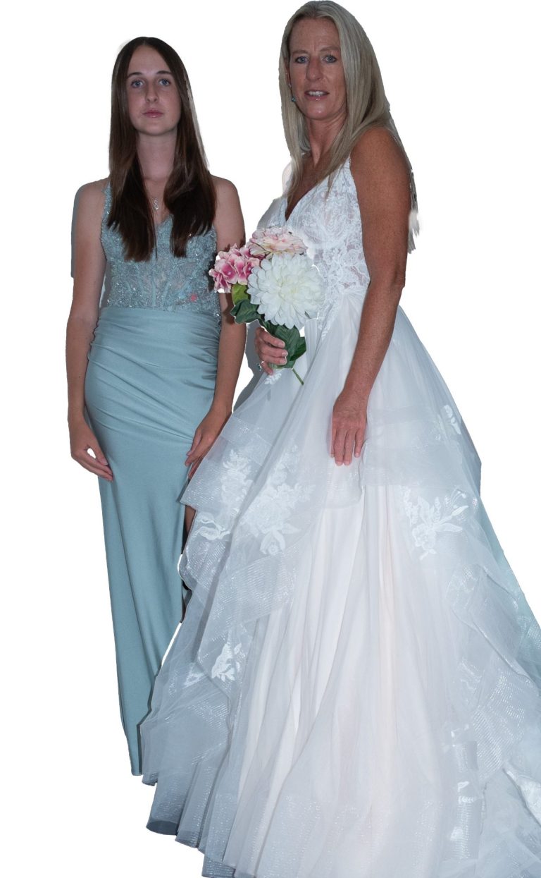 Two women stand together; one in a long blue dress, the other in a white wedding gown.