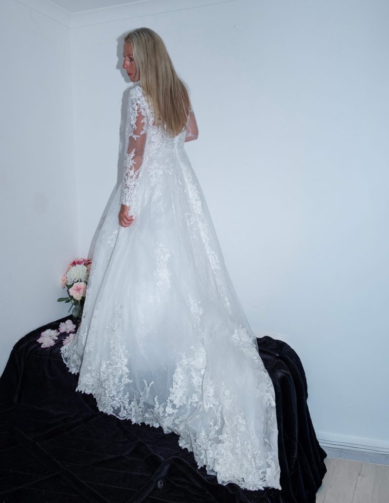 A woman in a white lace wedding dress, standing on a black surface with flowers nearby.