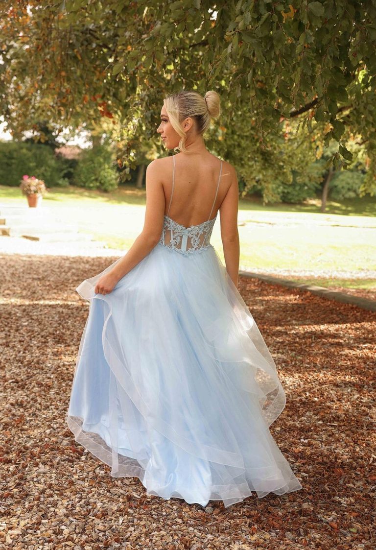 A woman in a flowing light blue gown stands with her back to the camera, outdoors.