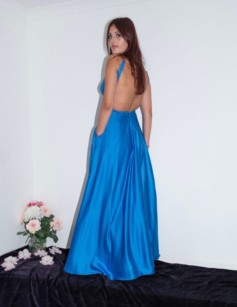 A woman in a blue gown with an open back, standing near flowers.