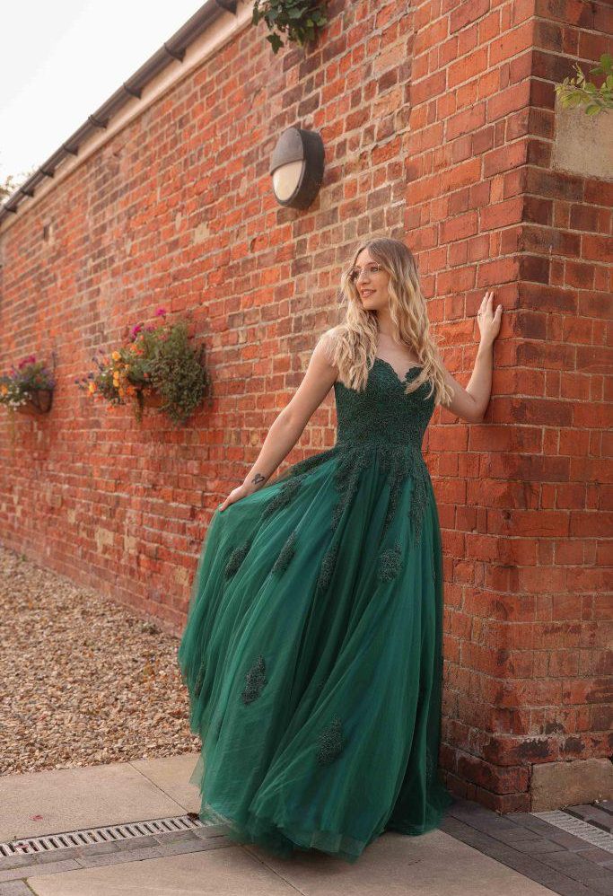 Woman in a green gown stands against a red brick wall, with flowers in the background.