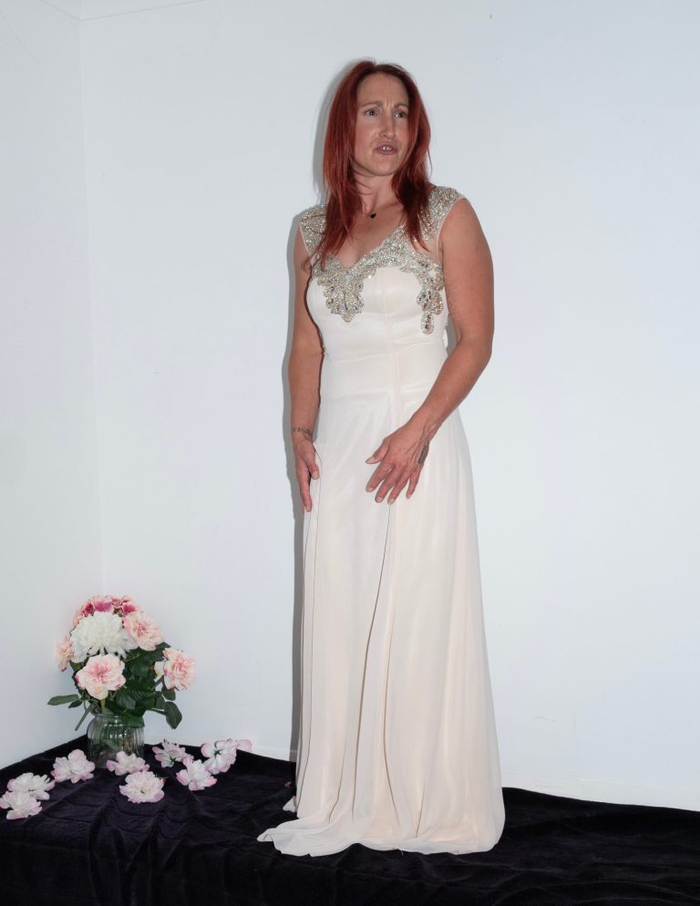 Woman in a light-coloured evening gown, standing beside a bouquet of flowers.