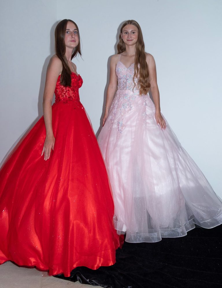 Two women wearing elegant gowns: one in red and one in pink, posed against a neutral backdrop.