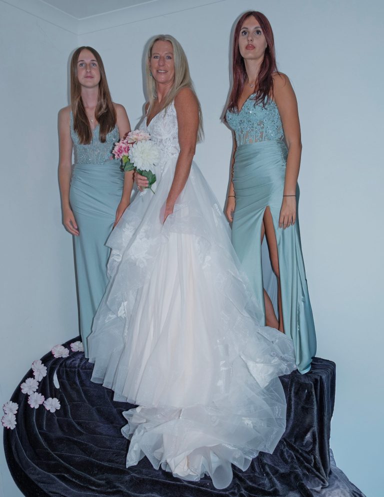 Three women in elegant dresses, one in a white bridal gown, posing with flowers.