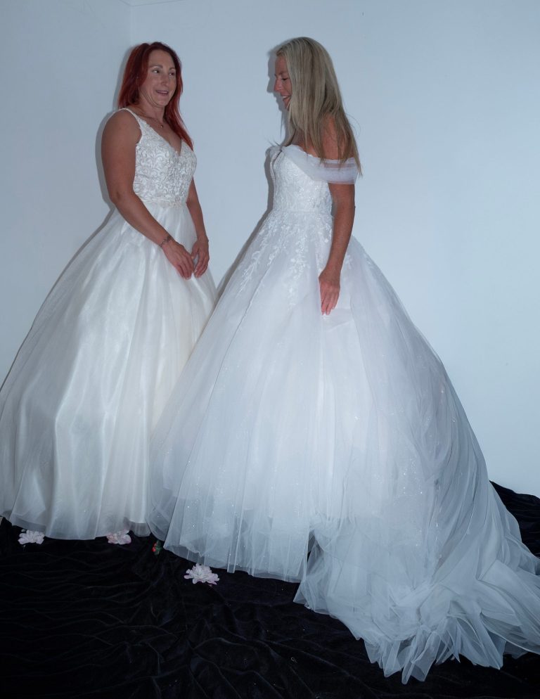 Two brides in elegant white wedding dresses standing together, smiling at each other.