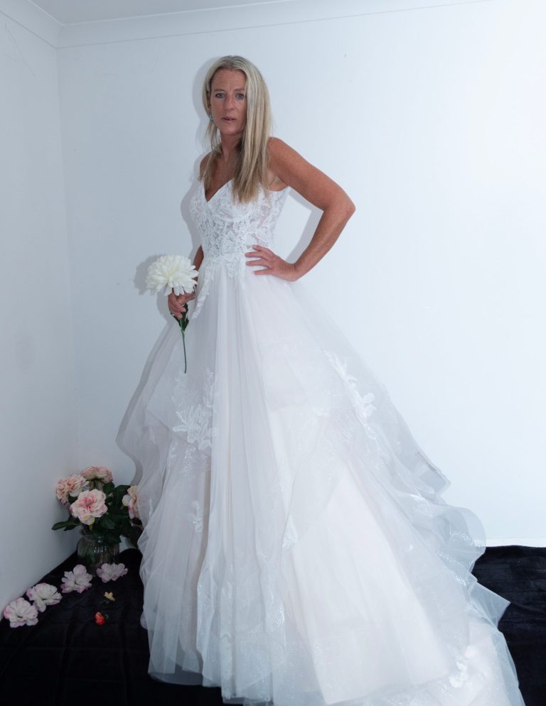Woman in a white wedding dress holding a bouquet, standing against a plain background.
