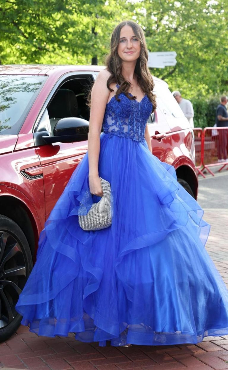 Woman in a blue ball gown holding a silver clutch, standing near a red car.