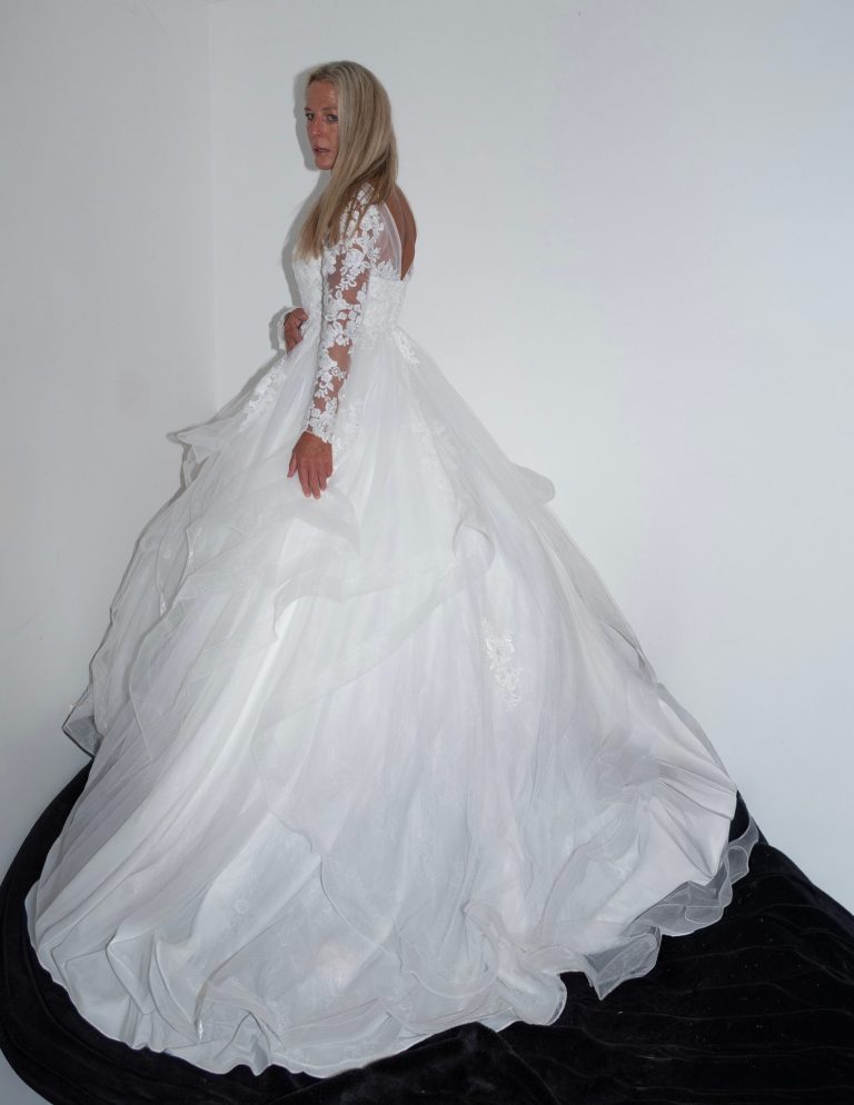 A woman in a flowing white wedding dress stands elegantly against a white background.