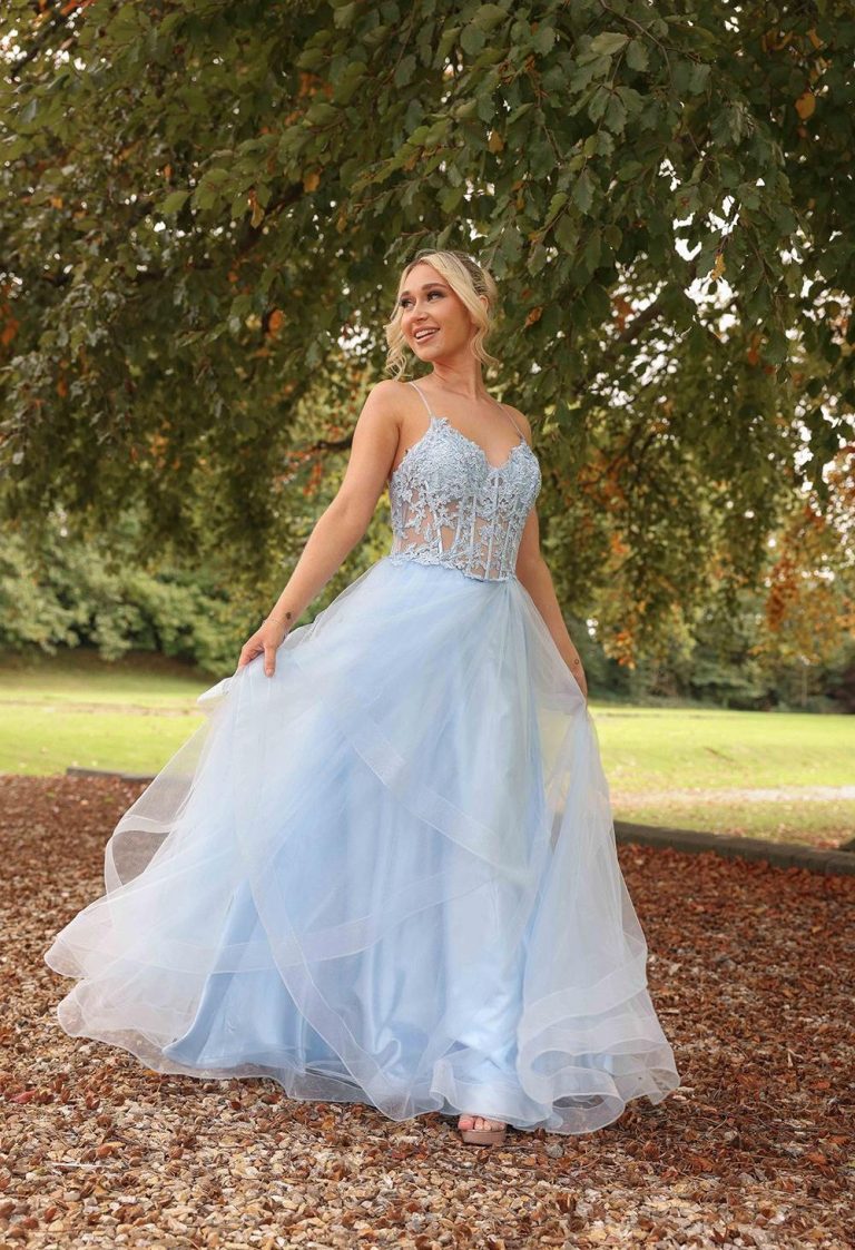 A woman in a light blue gown poses gracefully outdoors among trees.