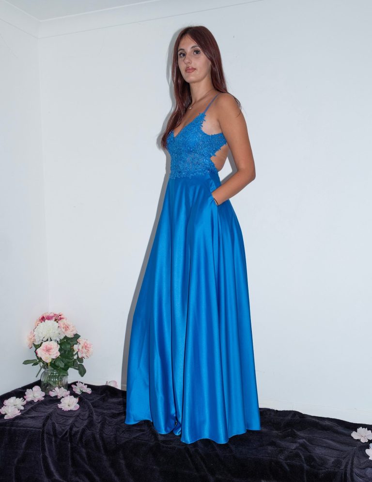 A woman in a long blue dress stands against a white wall, with pink roses nearby.