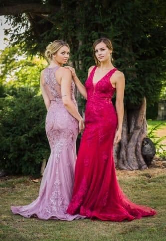 Two women in elegant evening gowns, one in pink and the other in red, pose outdoors.