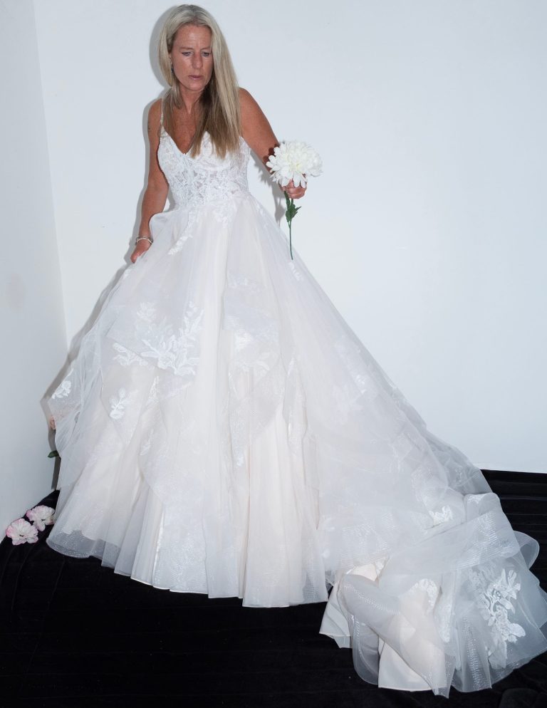 Woman wearing a white wedding gown, holding a flower, standing against a plain backdrop.