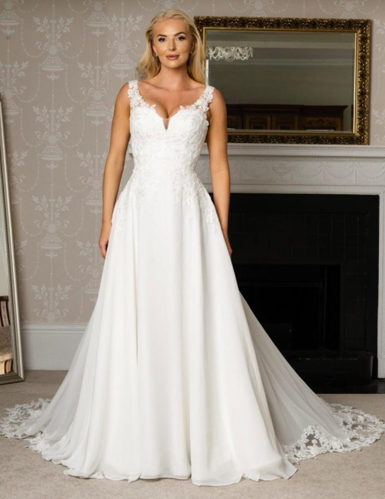 Woman in a silver-embellished white gown standing beside a floral arrangement.