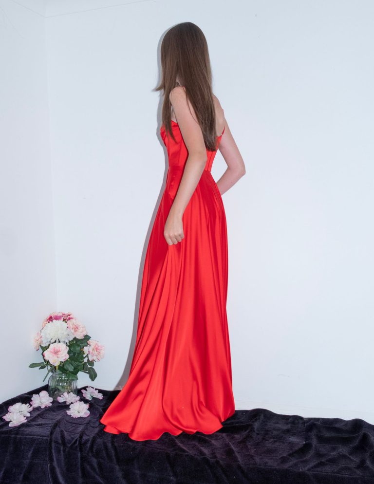 A woman in a flowing red dress stands with her back to the viewer, near pink flowers.