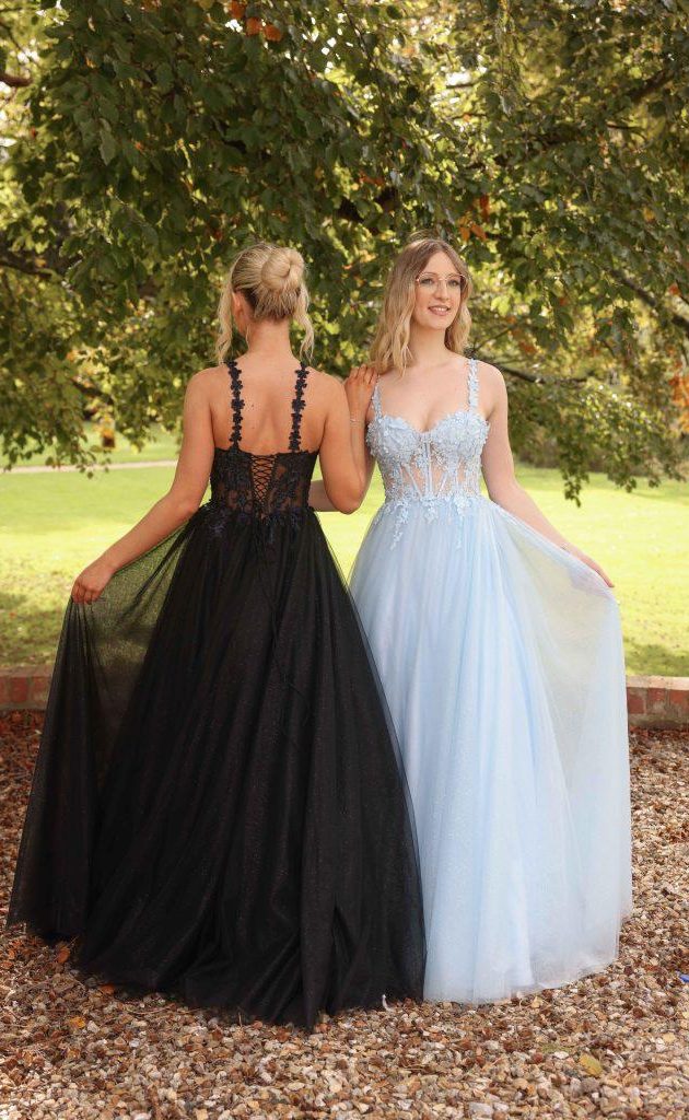 Two women in elegant gowns, one black and one light blue, standing back-to-back outdoors.